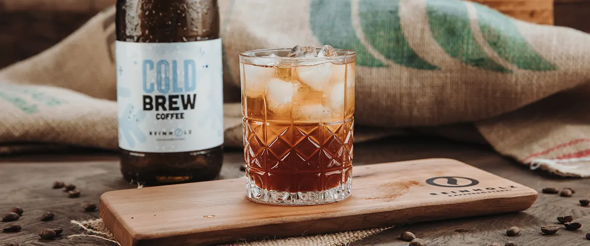 cold-brew-selber-machen-reinholz-eiskaffee Ein Glas Cold Brew Kaffee mit Eiswürfeln auf einem Holzbrett in der Reinholz Kaffeerösterei.