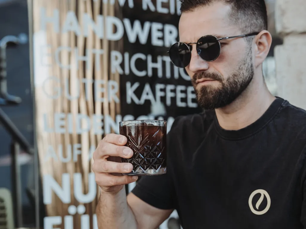 Reinholz-Barista hält ein Glas mit kaltem Kaffee vor dem Reinholz Kaffeeladen in Fulda.