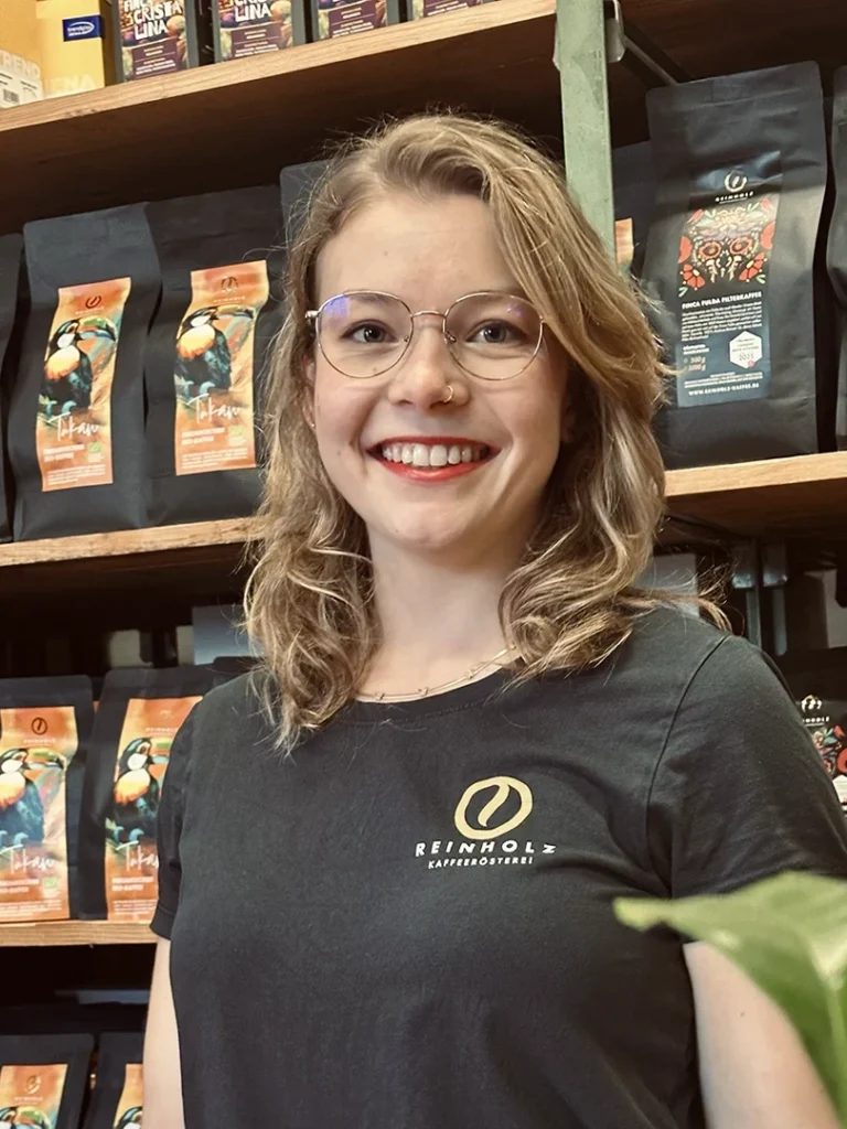 Reinholz-Praktikantin und Gastautorin Lisarah lächelt vor einem Regal mit Kaffeepackungen in der Reinholz Rösterei.